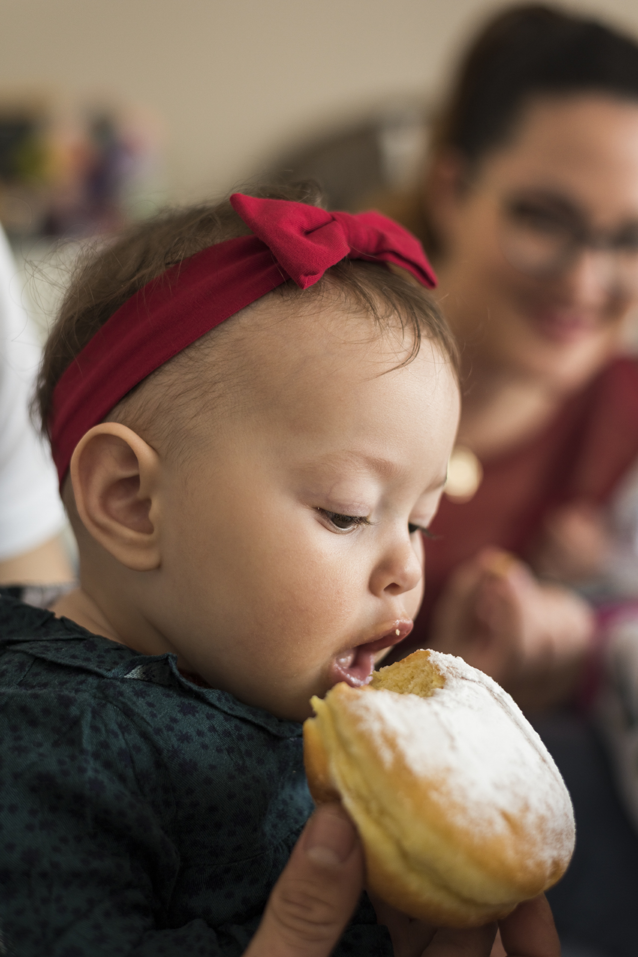 Bambina con fiocco rosso mentre mangia un dolce, con la madre sfocata sullo sfondo, progetto Storie di famiglia a Padova